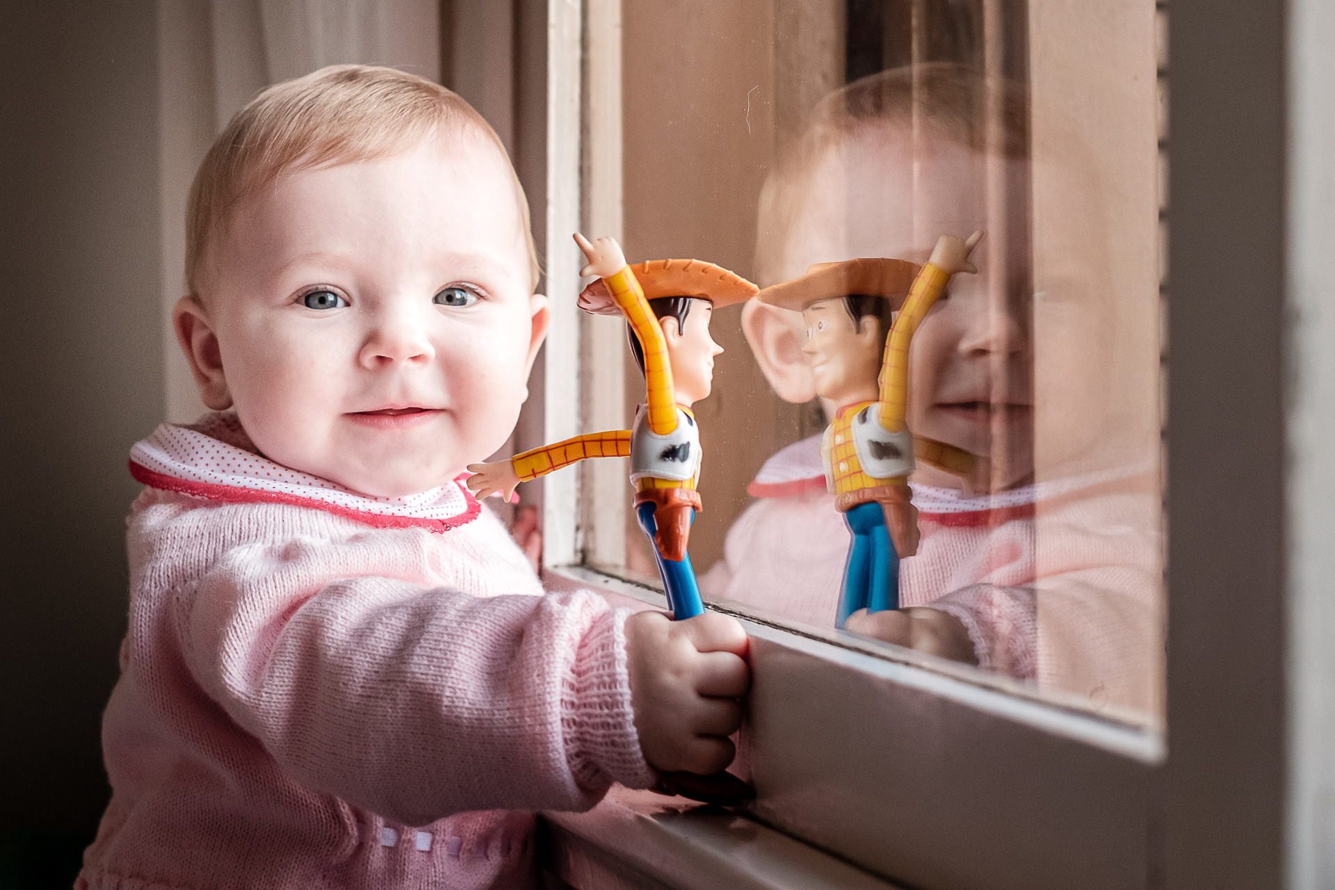Bebê junto à janela segurando um boneco Woody, entre reflexos no vidro, em fotografia de Kelly Schmidt