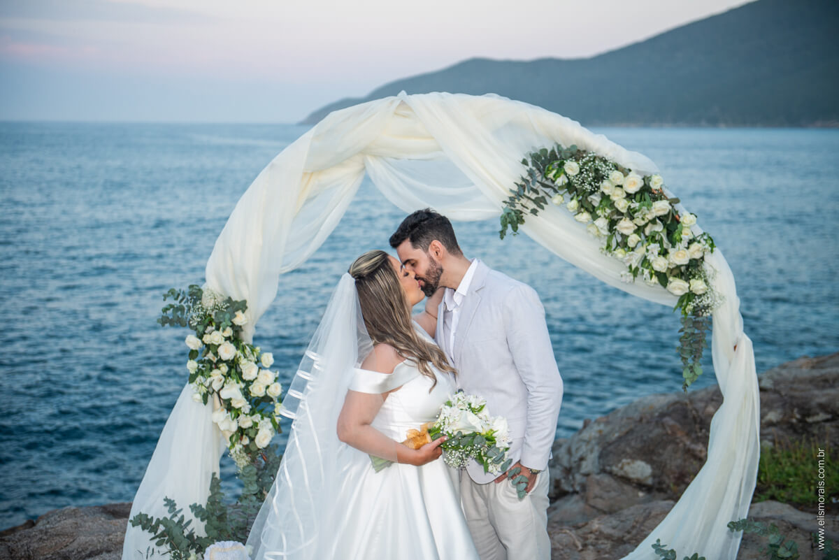 Noivos se beijando em frente ao cenário da decoração da cerimônia de casamento na praia no Pontal do Atalaia em Arraial do Cabo RJ. Elopement Wedding - Cerimônia de Casamento na Praia no Pontal do Atalaia em Arraial do Cabo RJ no Pontal da Grécia, Casa Mar da Grécia, Casa Grega, Santorini Terrace-Pontal do Atalaia, Casa Atenas / Greek House Arraial do Cabo RJ