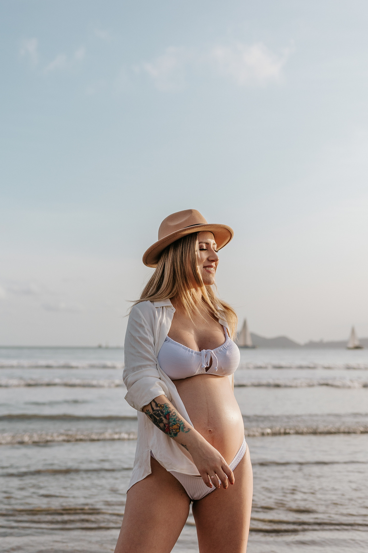Ensaio Gestante na praia de santos, grávida de biquini, camisa e chapéu com o por do sol e mar de fundo