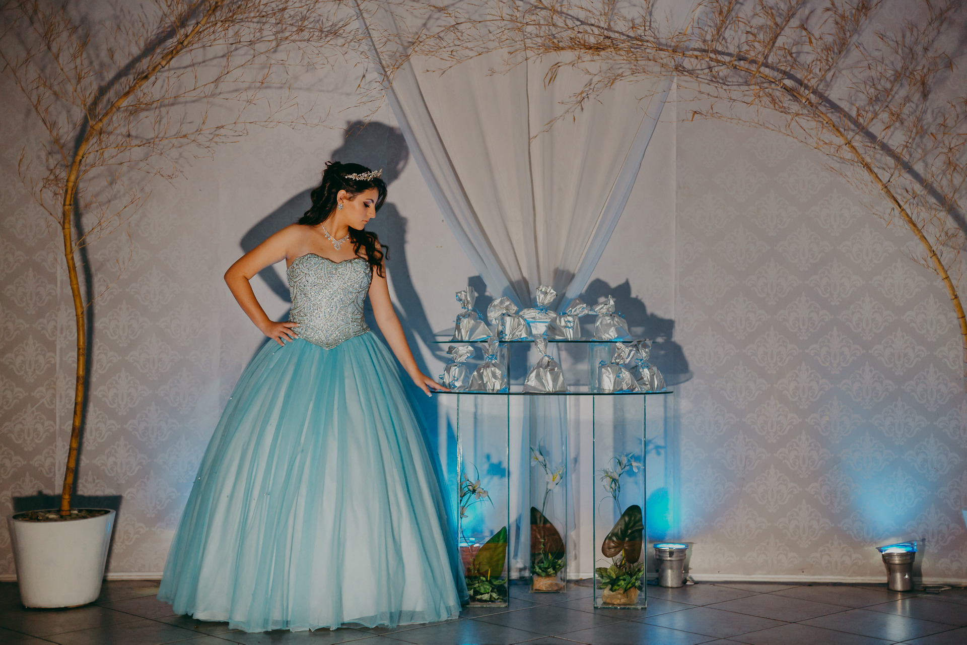 Fotografia de debutante com luz bem marcada em que olha para mesa de lembrancinhas numa pose elegante e com vestido azul fotos de 15 anos na SVM Festas em Piedade no Rio de Janeiro