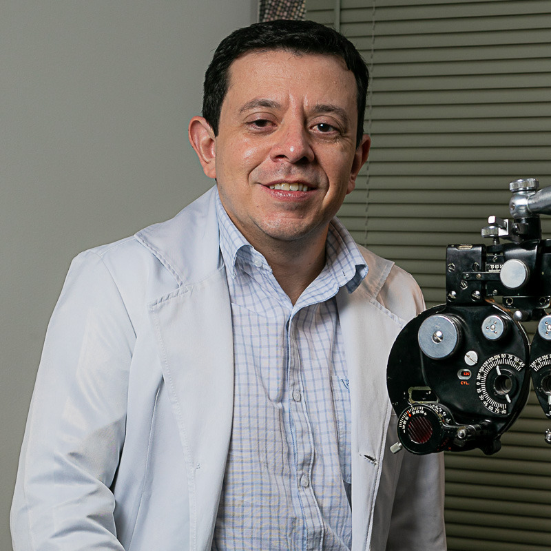 Homem, meia idade, sem barba, cabelos pretos curtos, vestindo jaleco branco, camisa listrada fina, no fundo parede branca com persiana fechada e equipamento oftalmológico para verificar o grau, ou teste de visão