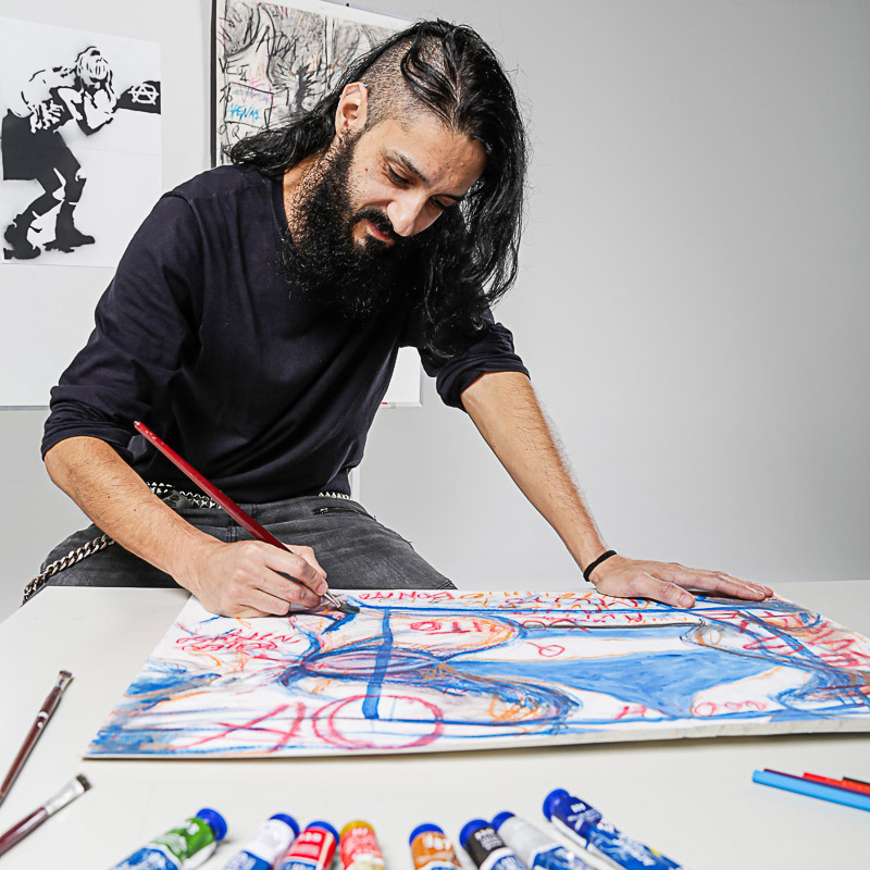 Homem, adulto, com cabelos longos e raspado acima da orelha, sentado em banco mais alto que a mesa, apoiado as mãos, fazendo um desenho colorido, ao fundo neutro com alguns desenhos autorais em cor monocromático, e na mesa alguns acessórios coloridos para sua arte.