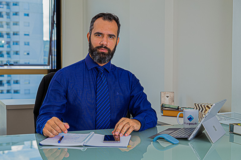 Homem, cabelos curtos, barba com fechando o rosto, camisa azul, papelaria de escritório, computador portátil, janela ao fundo e vista externa de prédios com parede de cor neura.