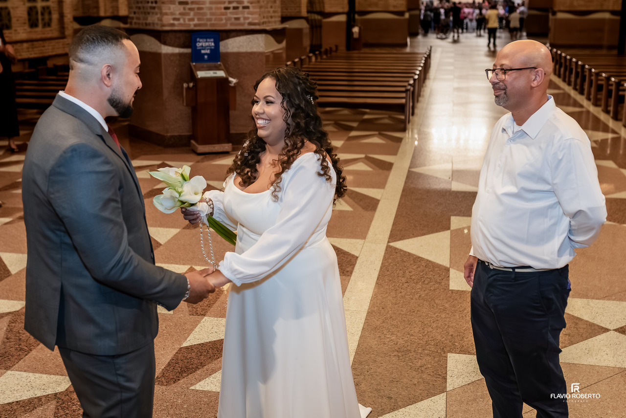 Noiva e noivo sorrindo durante cerimônia de casamento no Santuário Nacional de Aparecida com troca de alianças e buquê branco