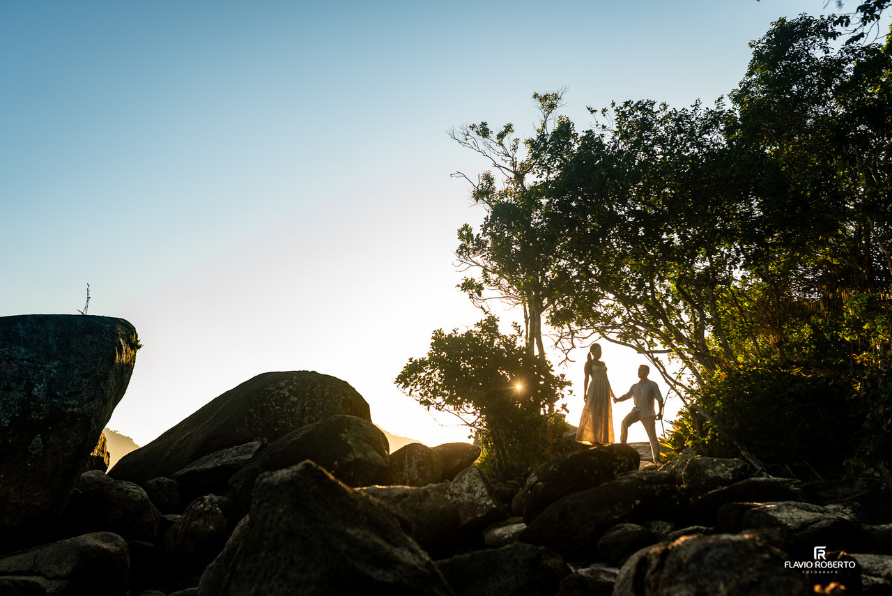 Casal em silhueta no topo das pedras em Ubatuba durante o pôr do sol, com luz dourada atravessando as árvores e criando efeito artístico.
