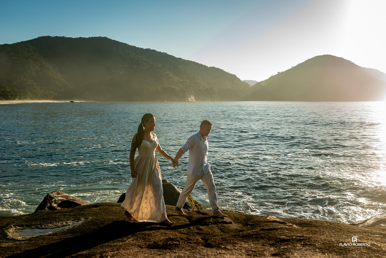 Casal caminhando de mãos dadas sobre pedras à beira-mar em Ubatuba durante ensaio pré-wedding ao pôr do sol.