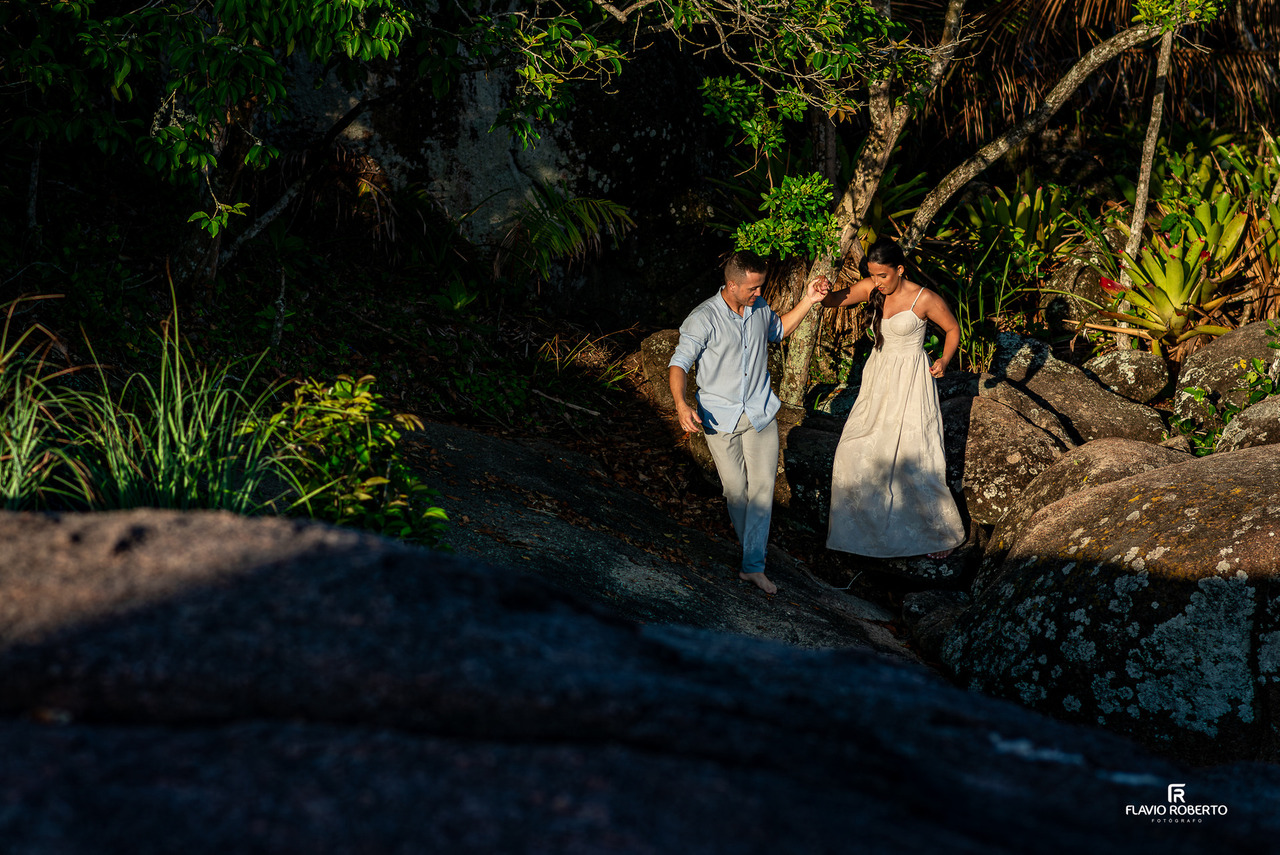 Casal de noivos em ensaio pré-wedding em Ubatuba caminhando sobre pedras com árvores e luz natural suave ao fundo.