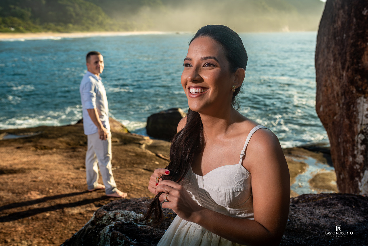 Retrato da noiva sorrindo em primeiro plano durante ensaio pré-wedding em Ubatuba, com o noivo ao fundo e o mar ao redor.