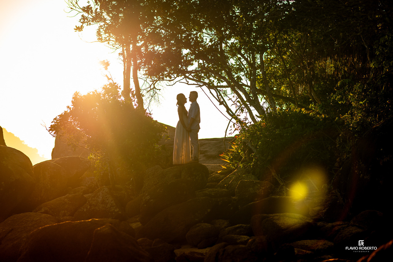Casal de noivos em ensaio pré-wedding em Ubatuba abraçado sobre pedras com árvores e luz natural suave ao fundo.