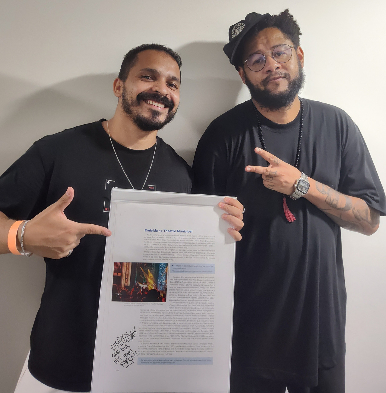 Fotógrafo Amauri Rafael ao lado do cantor Emicida, segurando uma página de livro didático que exibe sua fotografia autoral e o autografo do proprio emicida no qual ele escreveu "que dia  ein meu parça?!"