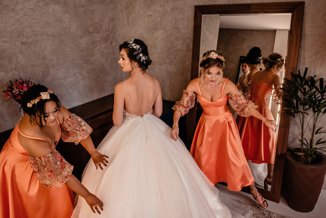 Madrinhas de vestido laranja ajudam a noiva a compor o vestido de noiva ao espelho, preparativos do casamento