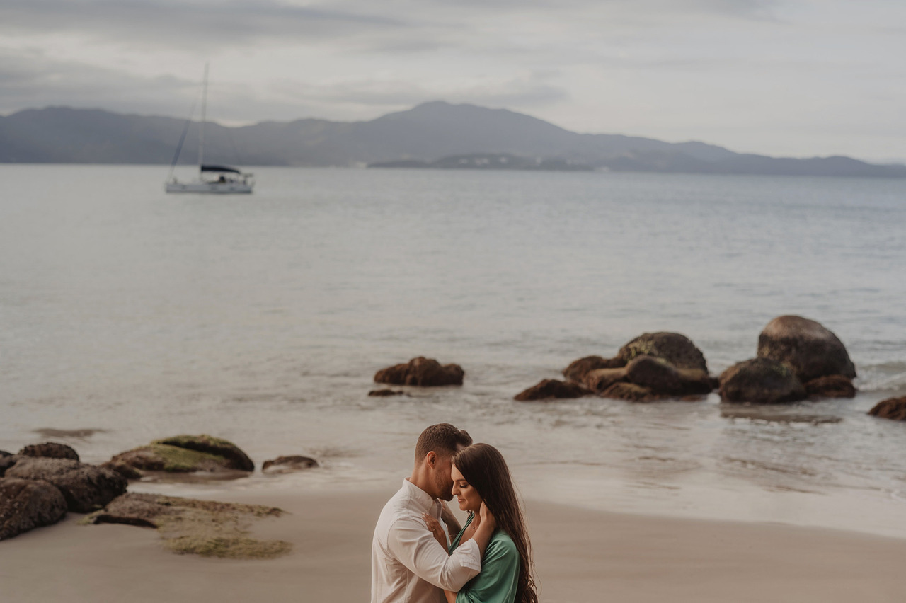 Casal abraçado numa praia com rochas e um veleiro ao fundo, sessão de noivos meses antes do casamento