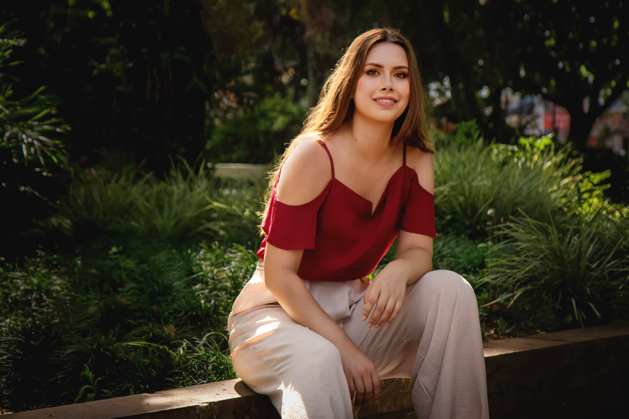 ensaio feminino-ensaio casual-ensaio fotografico em porto alegre-ensaio feminino em ivoti-ensaio feminino em dois irmaos-ensaio feminino na serra gaucha-garota jovem sentada em um muro baixo em uma praca-mulher jovem sorrindo vestindo uma blusa vermelha e calca longa rosa