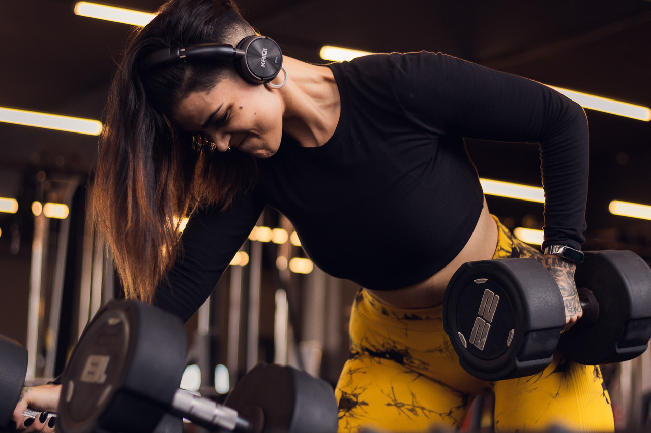 Ensaio feminino de uma mulher fazendo musculação, ela está inclinada segurando um halter com uma mão e com uma expressão de força e veias saltando pelo pescoço, mostrando um esforço.