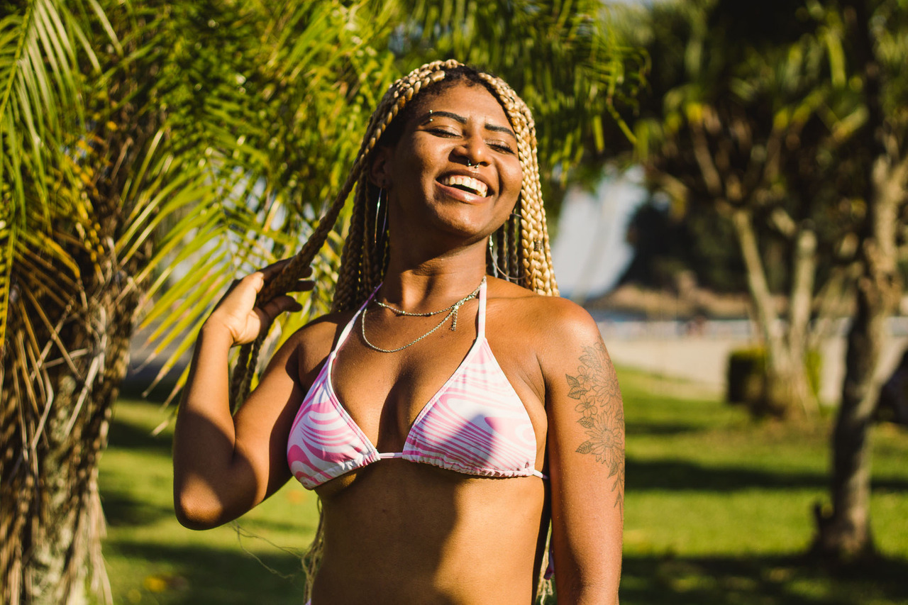 Ensaio feminino na praia, em santos, com o fundo de natureza com coqueiros, gramado verde e parte da areia e da água do mar, a modelo está sorrindo enquanto joga os cabelos para trás, com um sol lateral em seu corpo.
