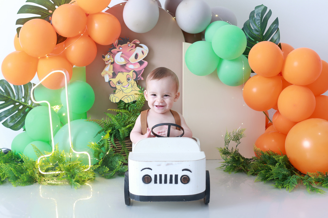 Bebê de 1 ano sorrindo dentro de um jipe ​​de brinquedo branco com luminoso de LED número um em cenários de balões laranja e verde no estúdio Jandira Ferreira Fotografia.