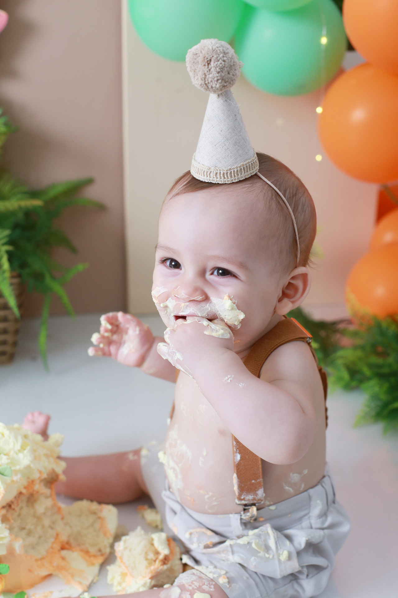 Close de bebê com chapéu de aniversário com pompom se lambuzando com bolo em ensaio Smash the Cake em São Paulo.