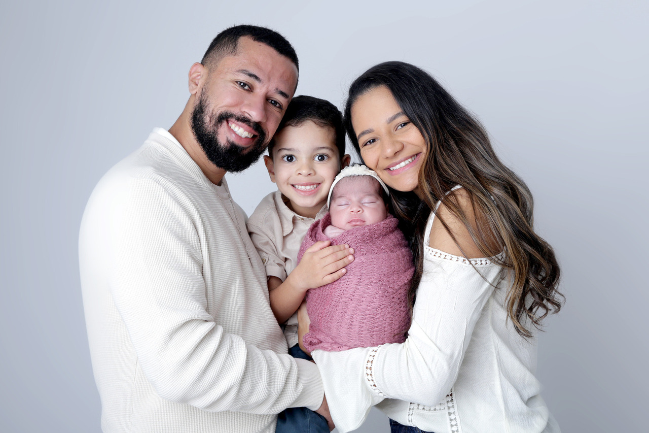Retrato de família em estúdio fotográfico com pai, mãe, filho mais velho sorrindo e bebê recém-nascida Helena envolvida em envoltório rosa, em São Paulo.