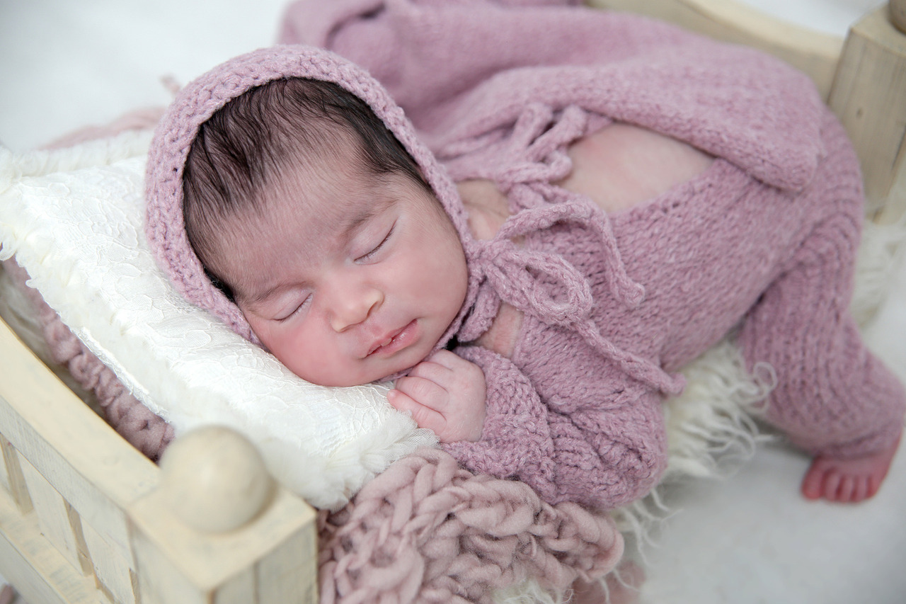 Bebê recém-nascida Helena dormindo de touca e roupinha de tricô rosa em mini cama de madeira no estúdio de fotografia newborn em Pirituba.