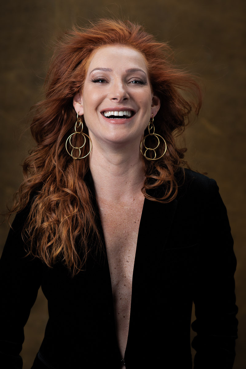 Retrato vibrante de mulher ruiva sorrindo com liberdade, usando blazer preto e brincos grandes. Ensaio feminino com presença marcante e luz controlada.