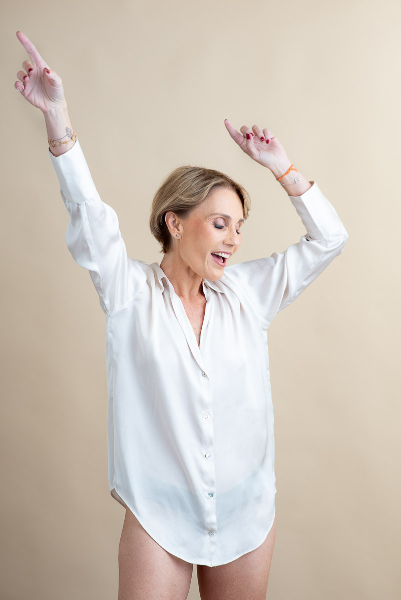 Retrato espontâneo de mulher sorrindo com alegria, vestindo camisa branca oversized. Ensaio feminino com luz suave e expressão autêntica.