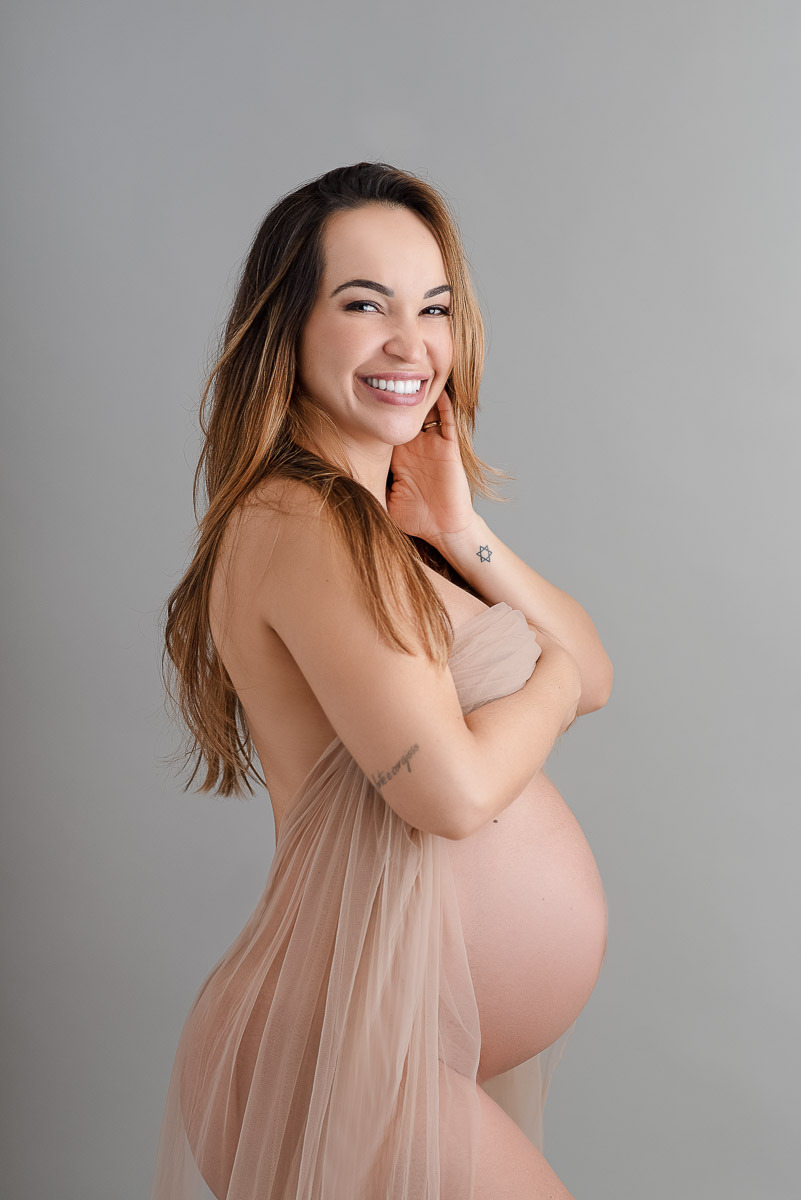 Retrato colorido de gestante com vestido fluido em tom nude, sorrindo com delicadeza — ensaio de maternidade com fundo neutro