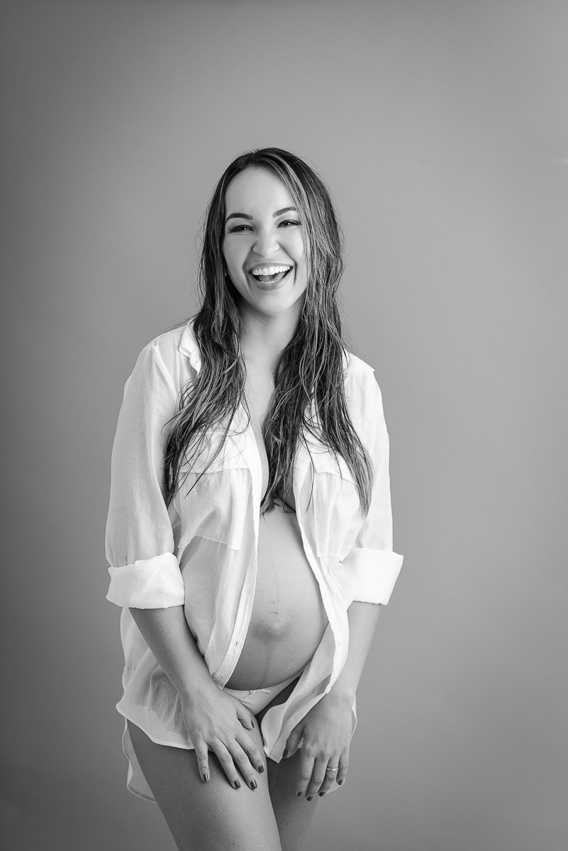 Retrato em estúdio de gestante sorridente em camisa branca, fotografia em preto e branco com estilo leve e natural