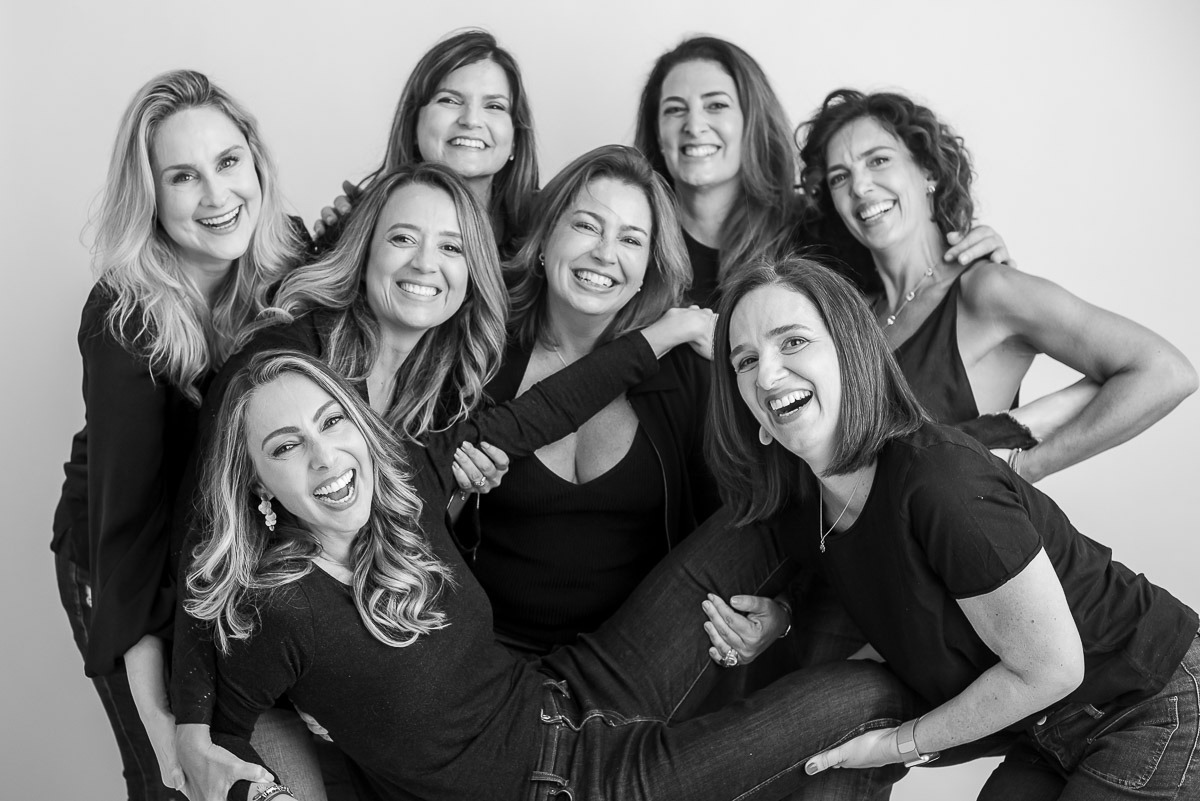 Retrato espontâneo de grupo de amigas rindo e abraçadas, em estúdio com fundo branco — ensaio coletivo feminino em preto e branco