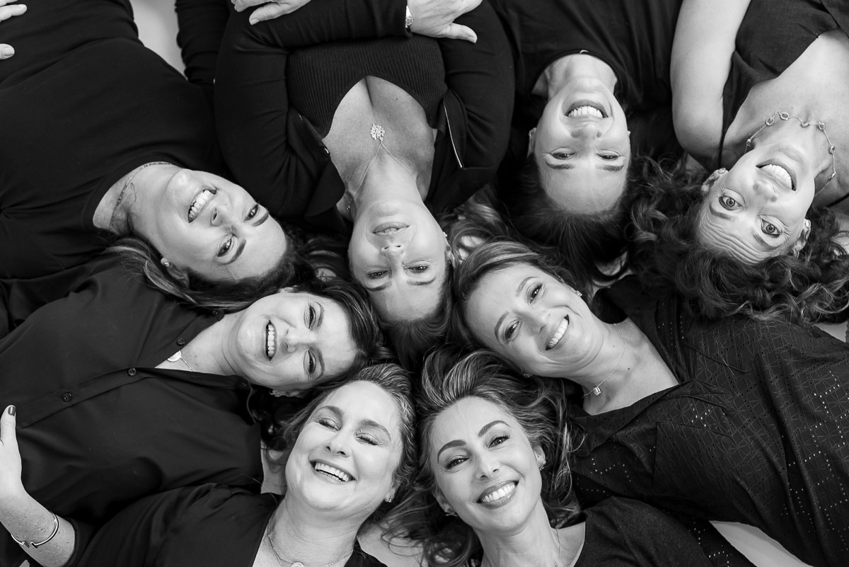 Retrato em grupo de mulheres maduras deitadas em círculo, sorrindo, em preto e branco — ensaio coletivo com clima afetuoso e divertido