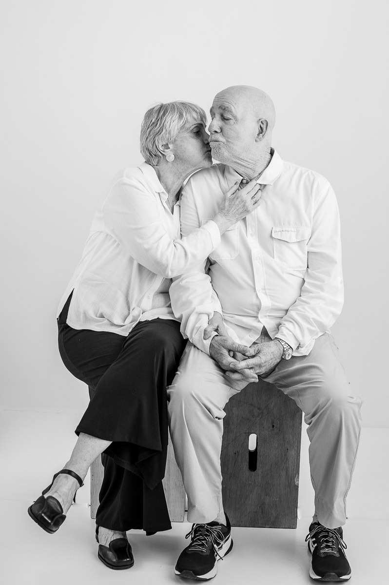 Retrato afetuoso de casal idoso em estúdio — mulher beija o companheiro sentados lado a lado, imagem divertida e carinhosa