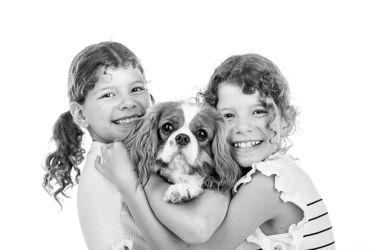 Retrato em estúdio de duas meninas sorridentes abraçando um cachorro — imagem afetiva e espontânea em preto e branco