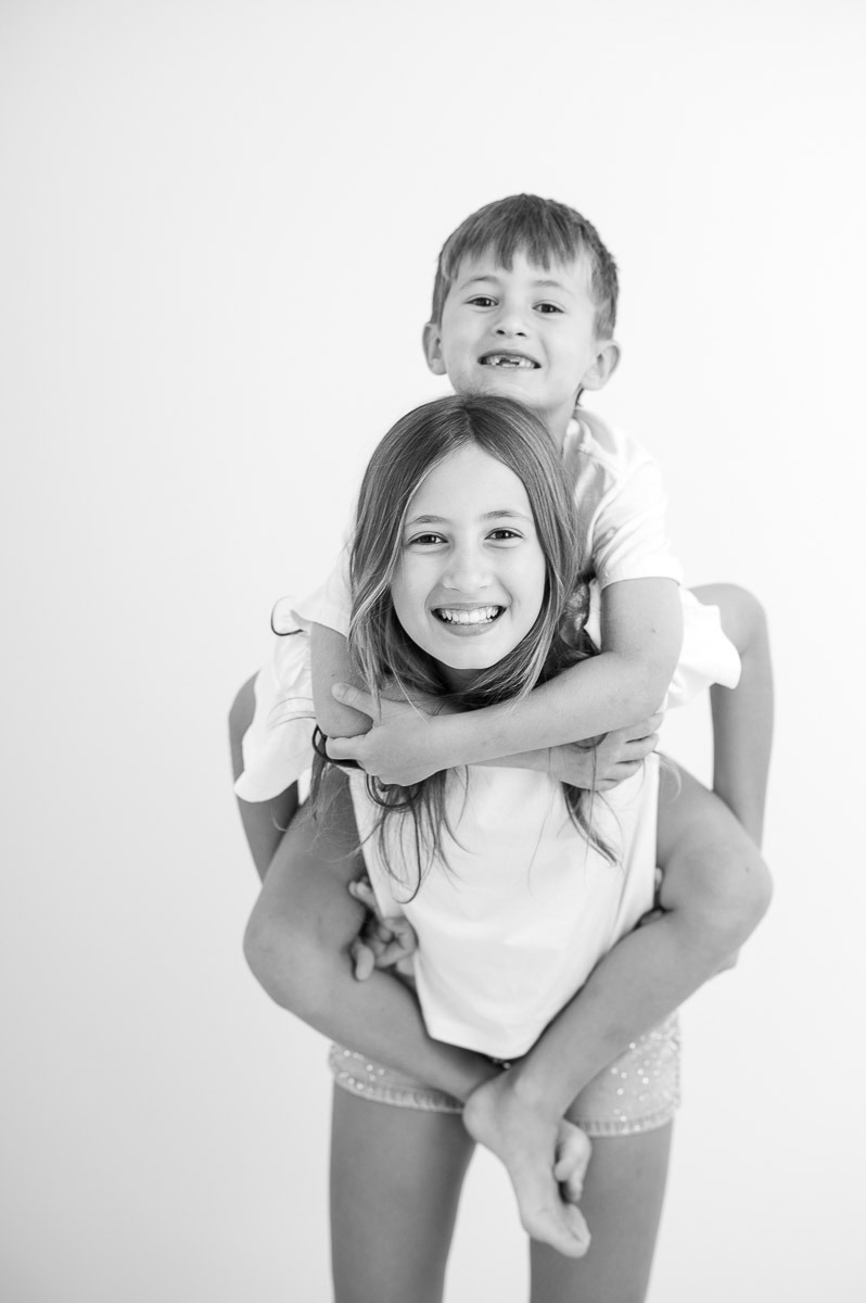 Retrato espontâneo de irmãos brincando em estúdio — menina carregando o irmão nas costas com expressão alegre