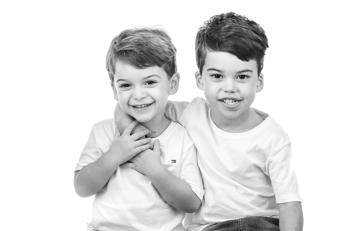 Retrato infantil em estúdio com dois irmãos sorrindo e abraçados — fotografia espontânea em preto e branco com fundo branco