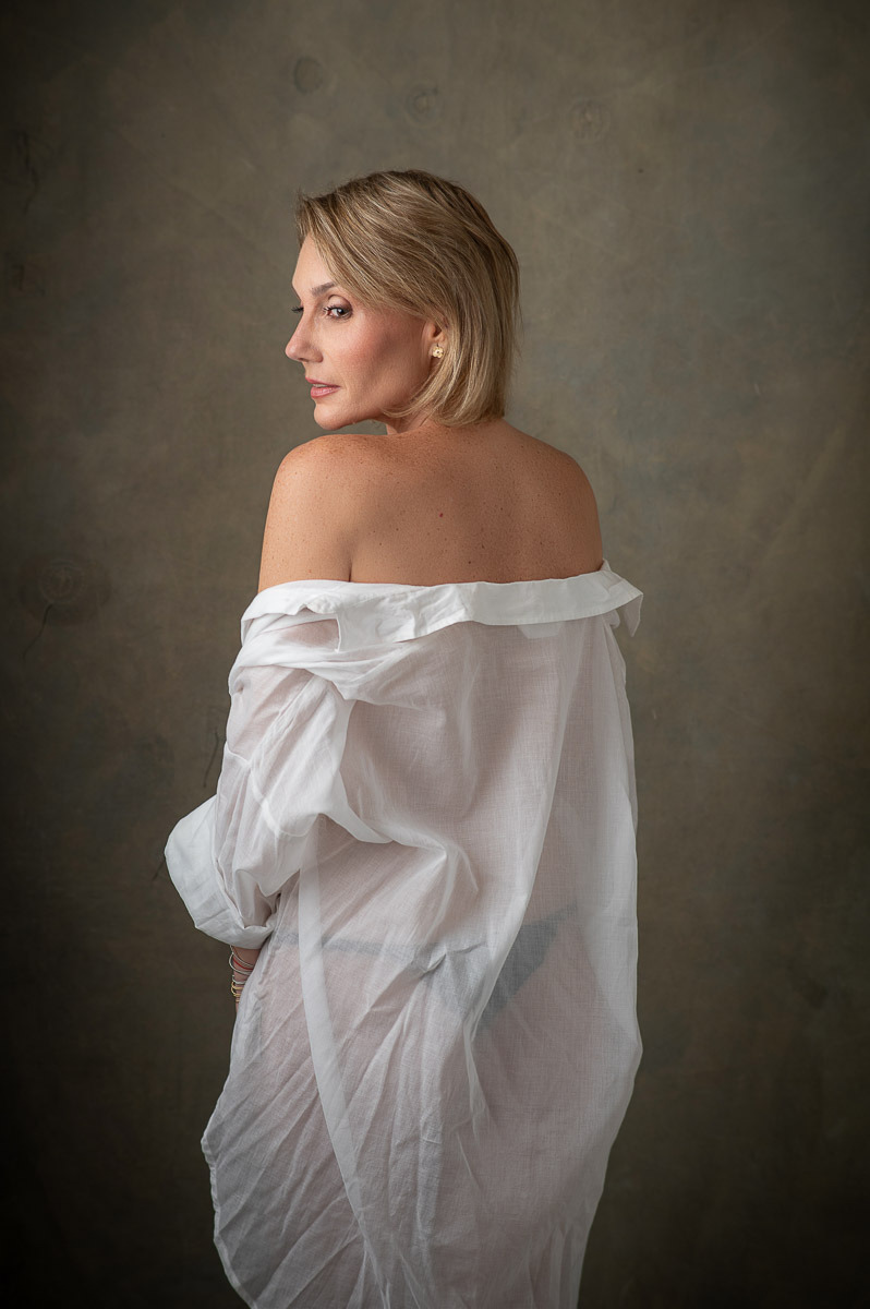 Mulher de costas parcialmente virada, usando camisa branca caída sobre os ombros. Retrato feminino delicado com atmosfera suave e minimalista.