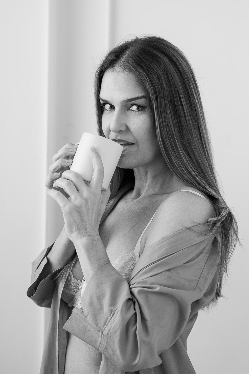 Mulher madura segurando uma caneca com olhar confiante e sedutor, vestindo camisa aberta sobre lingerie. Retrato sensual em preto e branco com atmosfera leve e intimista.