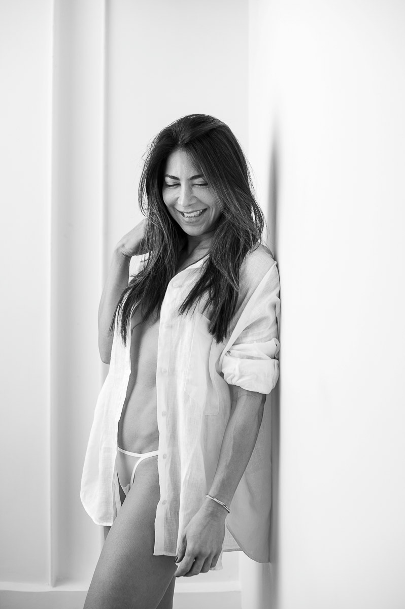 Mulher sorrindo com expressão espontânea e descontraída, vestindo camisa branca aberta sobre lingerie. Retrato em preto e branco com clima leve e autêntico.