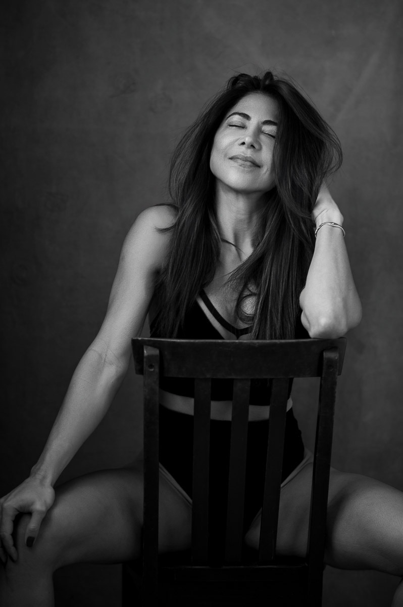 Mulher sentada de forma descontraída em uma cadeira, olhos fechados e expressão de prazer sereno. Ensaio sensual com atmosfera poética e intimista.