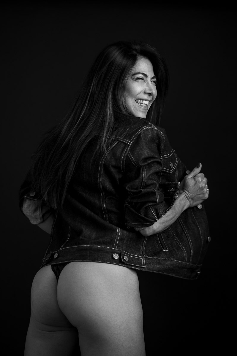 Mulher sorrindo espontaneamente, de costas, usando jaqueta jeans e lingerie. Fotografia em preto e branco com clima leve, confiante e divertido.