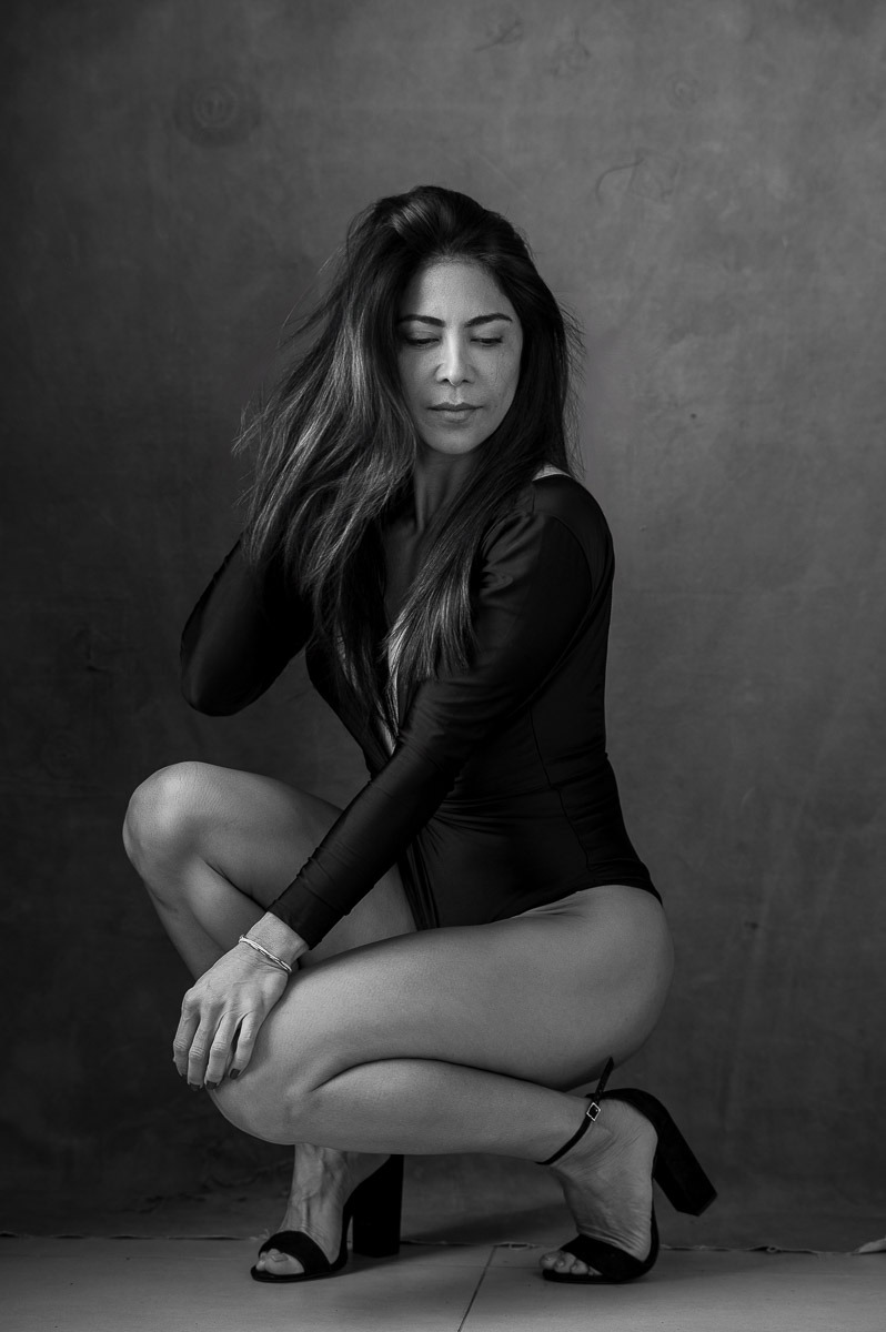 Mulher sentada com expressão introspectiva e postura elegante, usando body preto e salto alto, em um cenário minimalista. Retrato sensual com tom artístico e sofisticado.