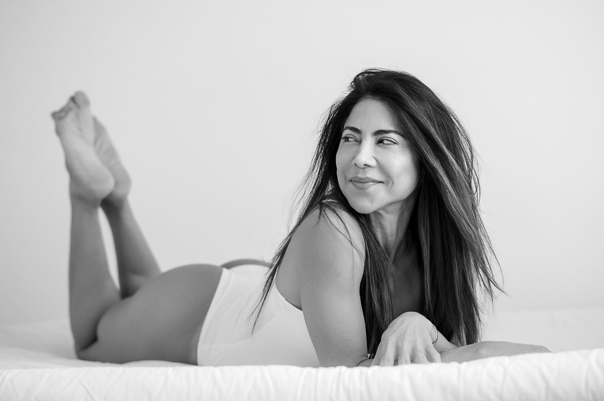 Mulher deitada de bruços na cama, olhando para o lado com expressão leve e relaxada. Ensaio sensual com luz natural e atmosfera acolhedora.