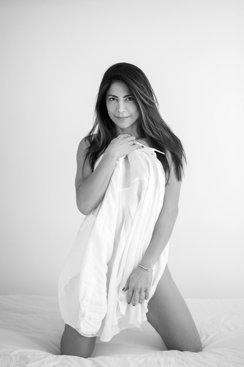 Mulher ajoelhada sobre a cama, segurando um lençol com leveza e olhar confiante. Retrato sensual com estética clean e acolhedora.