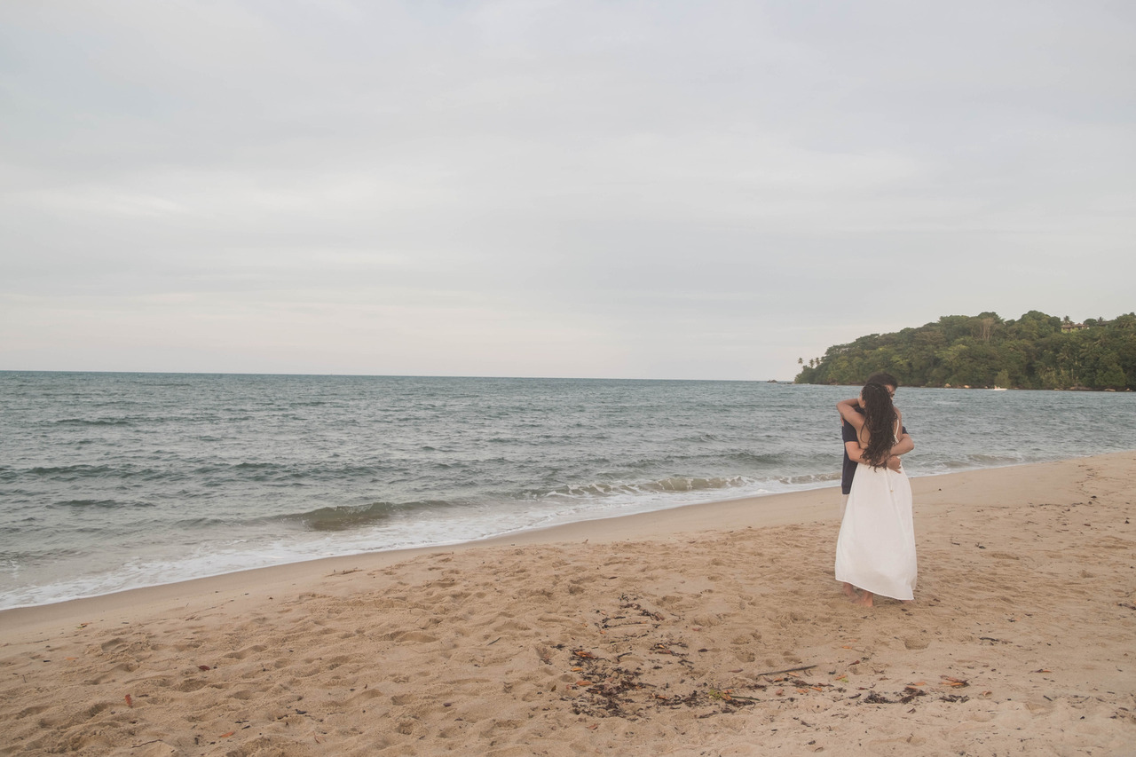 Leo e Lorrany se abraçam emocionados após um pedido de casamento surpresa durante um ensaio fotográfico em Boipeba, na Bahia, com o mar e o pôr do sol ao fundo.