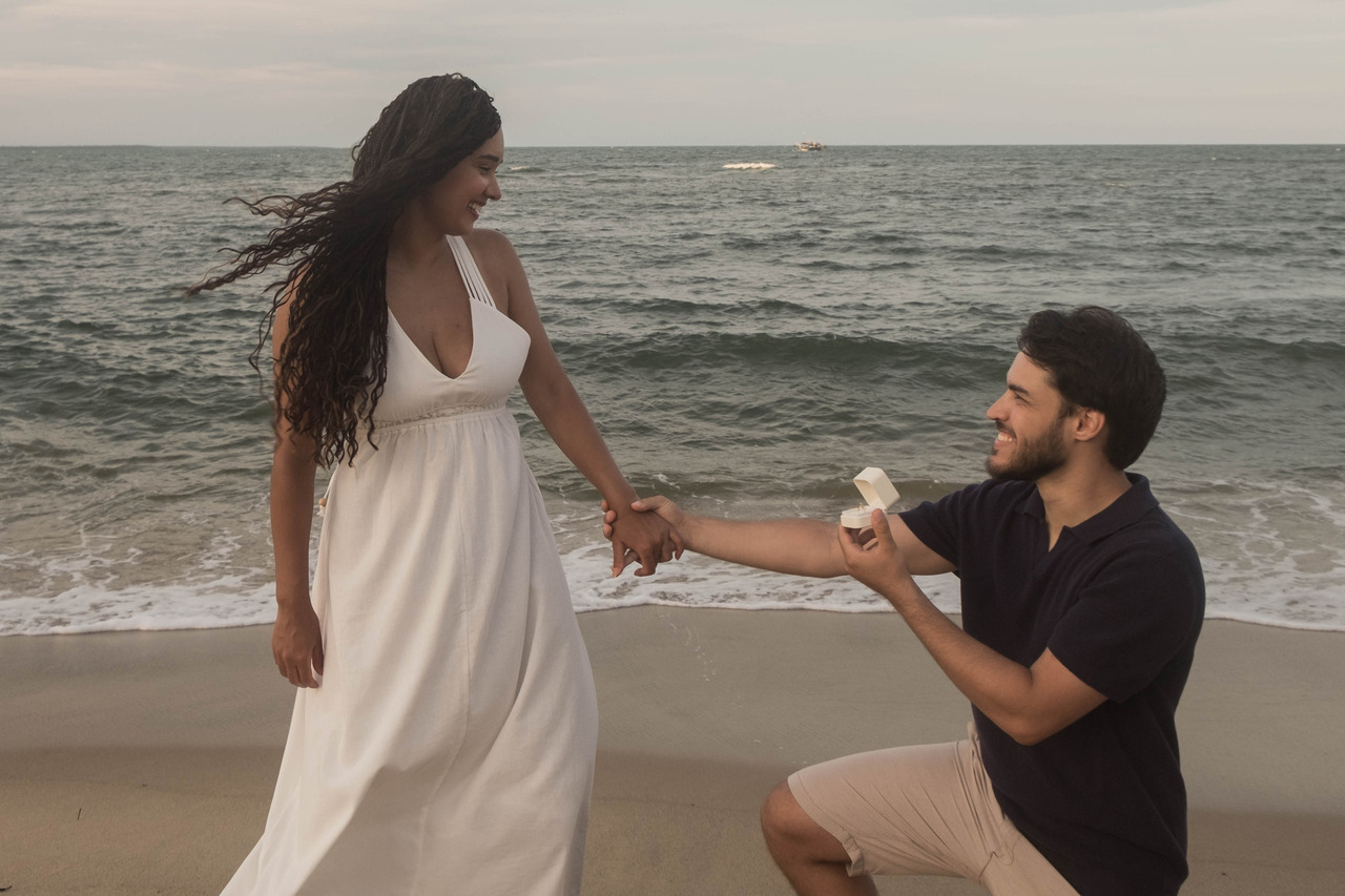 Leo ajoelhado fazendo um pedido de casamento surpresa para Lorrany durante um ensaio fotográfico ao pôr do sol na Praia da Boca da Barra, em Boipeba, Bahia.