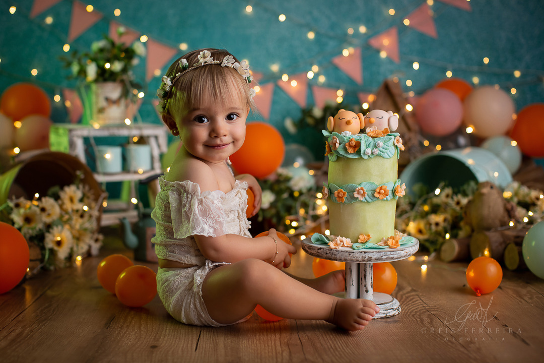 Smash the Fruit Jardim Encantado bolo de aniversário feito de melao para ensaio de bebe com passaros e flores de aplique de biscuit em um cenario fotografico com bebe de 11 meses sentada ao lado e luzes de pisca no fundo