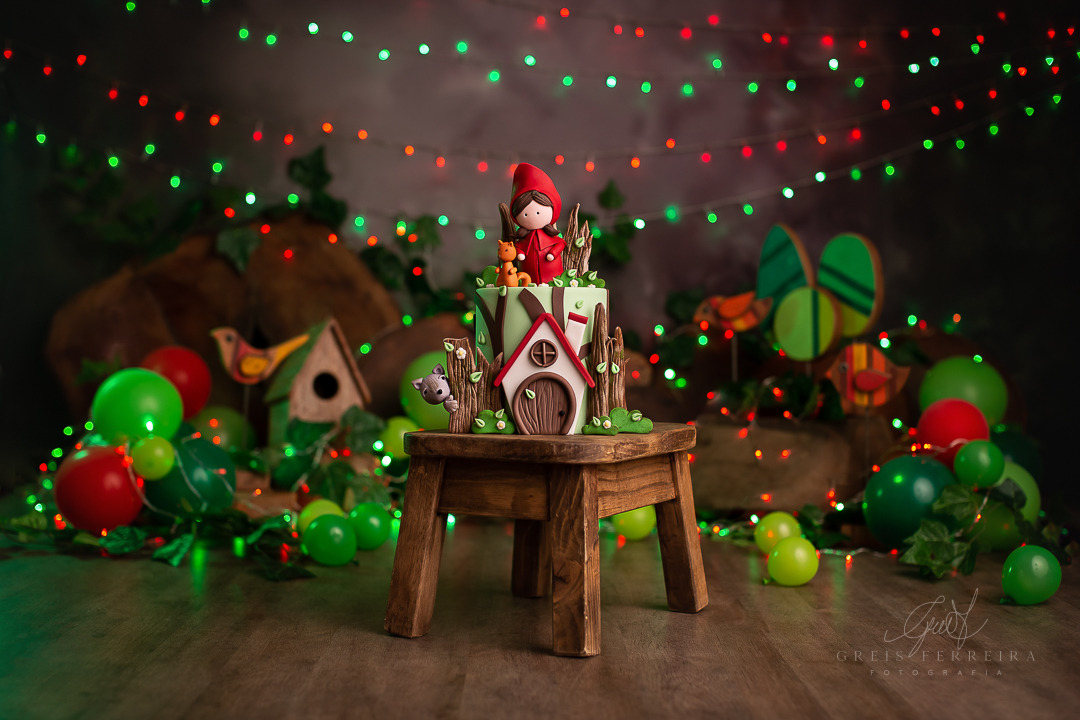 smash the cake chapeuzinho vermelho ensaio de bebe esmagando o bolo de aniversario com baloes, pisca-pisca verde e vermelho plantas e passatros de madeira ao fundo o bolo tem aparencia de uma casinha na floresta e no topo um biscuit da chapeuzinho cobde uma banquinho de madeira