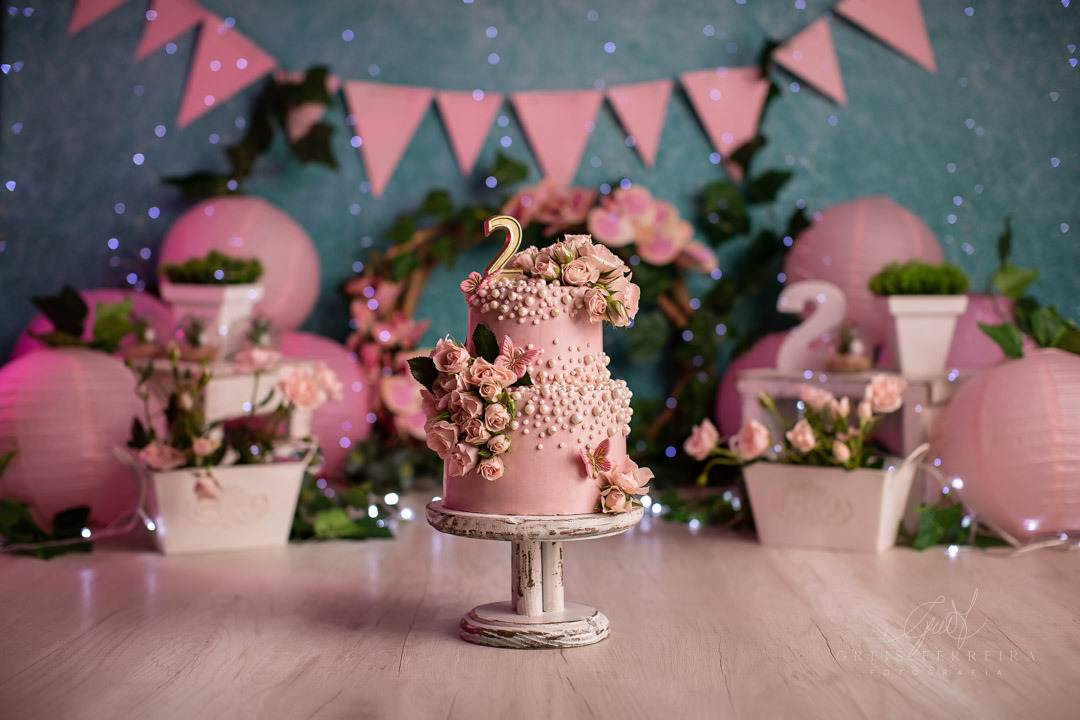 Smash the Cake Jardim Encantado com Flores ensaio de bebe tendo um bolo de aniversário a frente e com pisca ao fundo e cachepos de flores bandeirolas e baloes