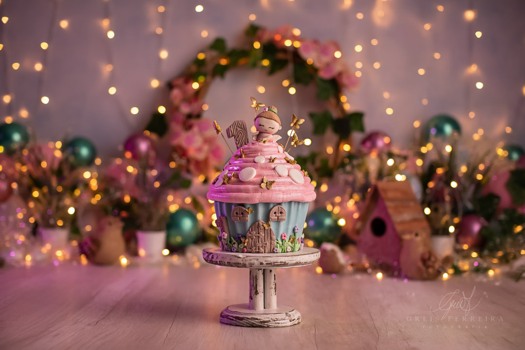 Smash the Cake Jardim Encantado com Fadas tendo um big cupcake a frente e um arco de flores com pisca ao fundo e casinhas de passarinhos e baloes