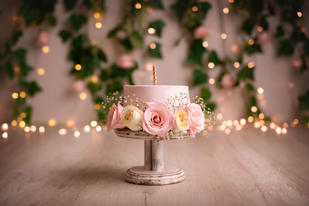Smash the Cake Jardim Encantado Clean com flores tendo um bolo de aniversário a frente e com pisca ao fundo junto com plantas e flores em uma madeira branca