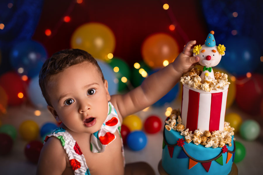 Smash the Cake ensaio bebe 1 ano tema palhacinho com bebe com maos em um bolo em formato de palhaco em cenario com tenda e baloes