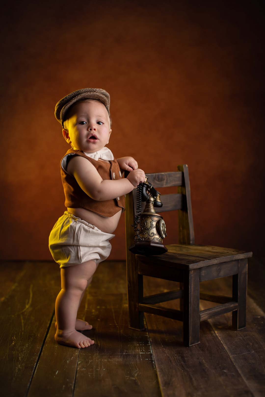 bebe usando ropa retro gravata boina e apoiando em uma cadeira de madeira e um telefone antigo ensaio de bebe